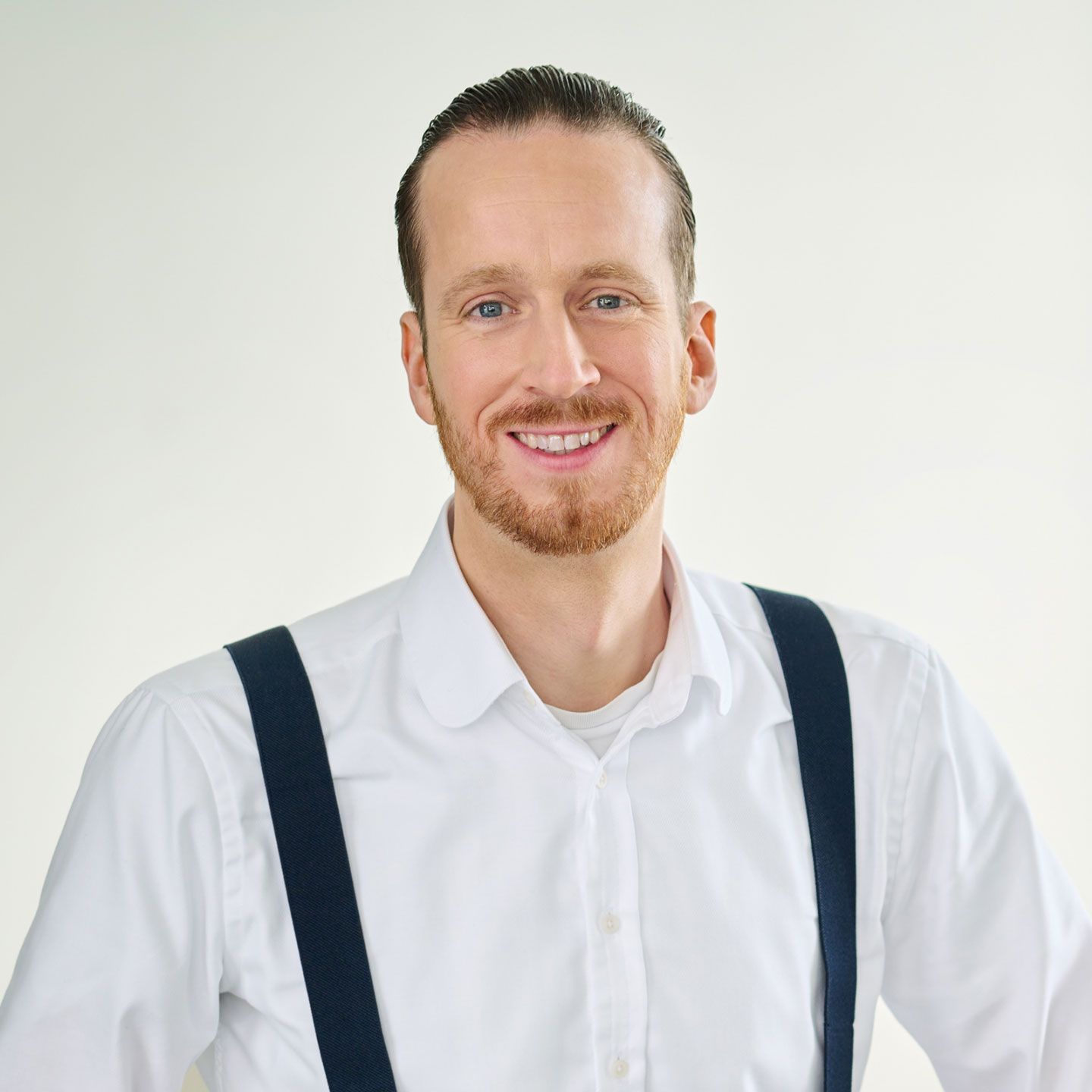 Sebastian Pollmer, mand står i hvid skjorte og blå seler i et lyst rum. 