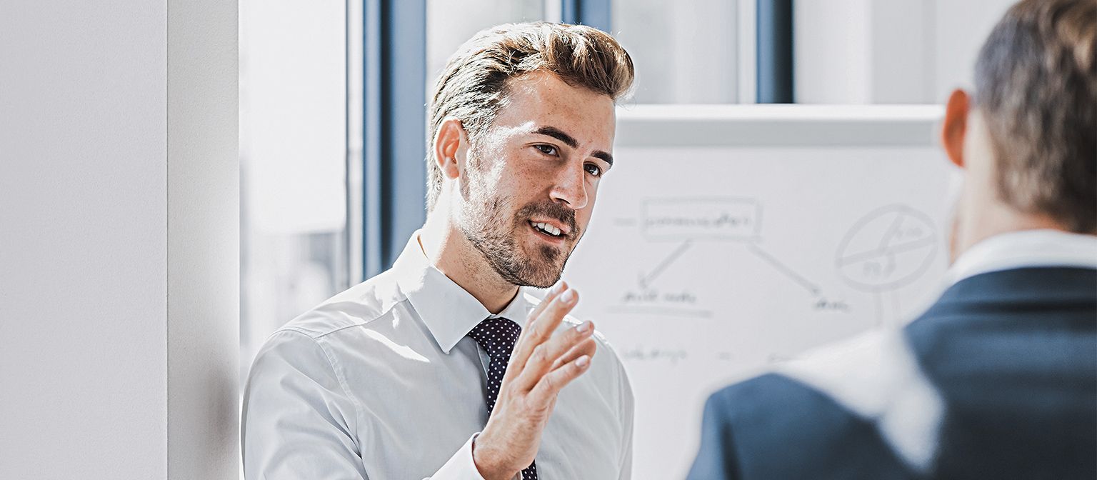 Man ser en mand i forretningstøj forklare noget med et venligt og engageret udtryk. Foran ham står en anden person, som tydeligt lytter til ham. I baggrunden ses en whiteboardtavle med en skitse.