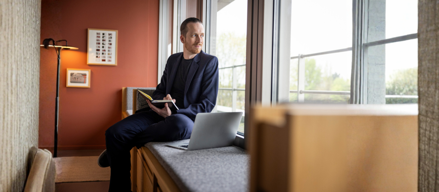 Sebastian Pollmer sidder i en vindueskarm med en åben laptop og en notesbog i hånden og stirrer ud ad vinduet.