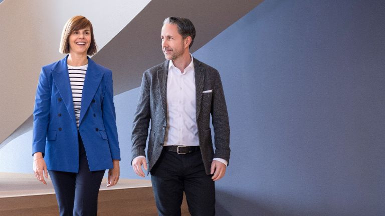 CEO'en i en grå blazer og CFO'en for EOS Gruppen i en blå blazer går gennem et moderne kontor. Rummet har store vinduer, en buet væg og stilfulde hængende lamper.