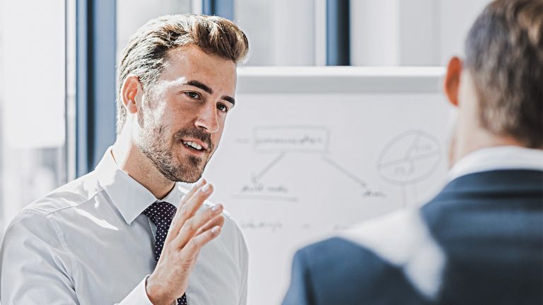 Man ser en mand i forretningstøj forklare noget med et venligt og engageret udtryk. Foran ham står en anden person, som tydeligt lytter til ham. I baggrunden ses en whiteboardtavle med en skitse.