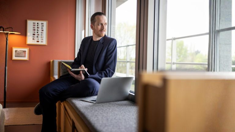 Sebastian Pollmer sidder i en vindueskarm med en åben laptop og en notesbog i hånden og stirrer ud ad vinduet.