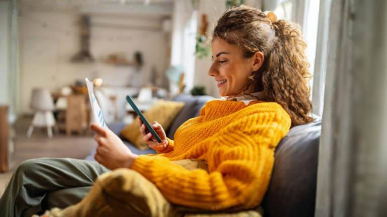 En smilende ung kvinde med krøllet hår, iført en hyggelig gul sweater, sidder i en sofa i sin stue og kigger på sin smartphone, mens hun holder et papirdokument.