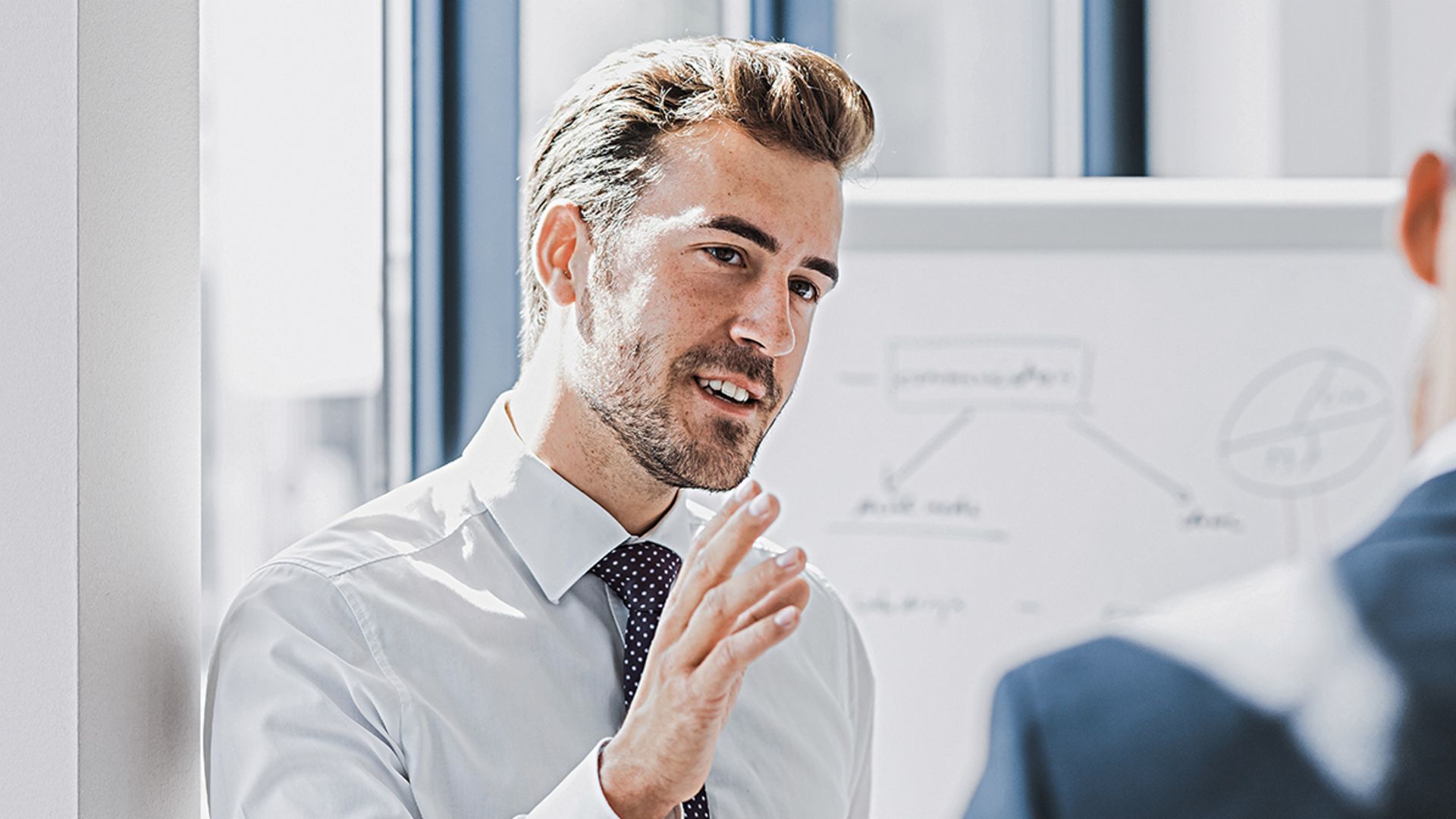 Man ser en mand i forretningstøj forklare noget med et venligt og engageret udtryk. Foran ham står en anden person, som tydeligt lytter til ham. I baggrunden ses en whiteboardtavle med en skitse.