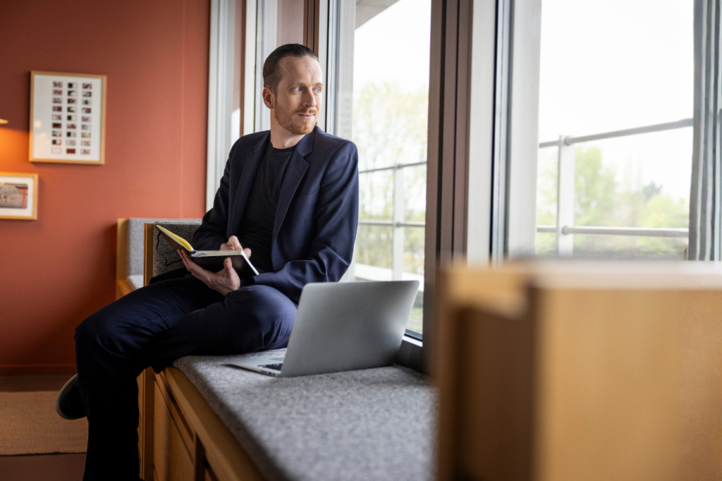 Sebastian Pollmer sidder i en vindueskarm med en åben laptop og en notesbog i hånden og stirrer ud ad vinduet.