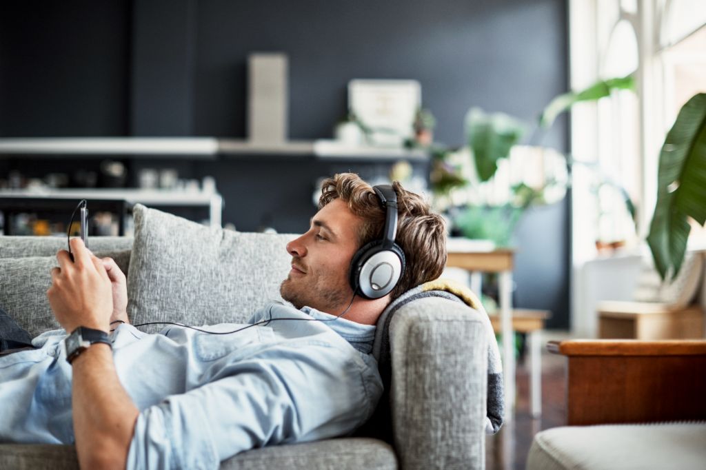 En ung mand ligger på en sofa i sin stue med hovedtelefoner på og kigger smilende på sin smartphone, mens han nyder musik eller en podcast.