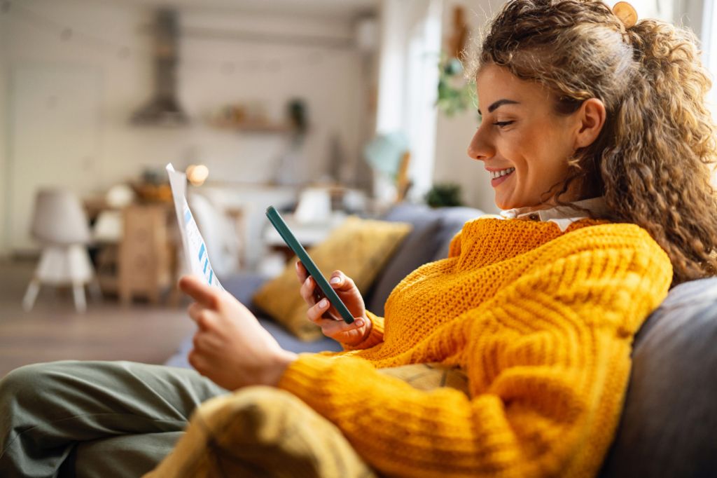 En smilende ung kvinde med krøllet hår, iført en hyggelig gul sweater, sidder i en sofa i sin stue og kigger på sin smartphone, mens hun holder et papirdokument.