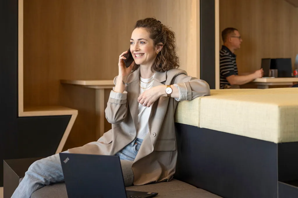 En smilende person sidder i en moderne kontorlounge, taler i telefon og arbejder på en bærbar computer. I baggrunden arbejder to andre personer.