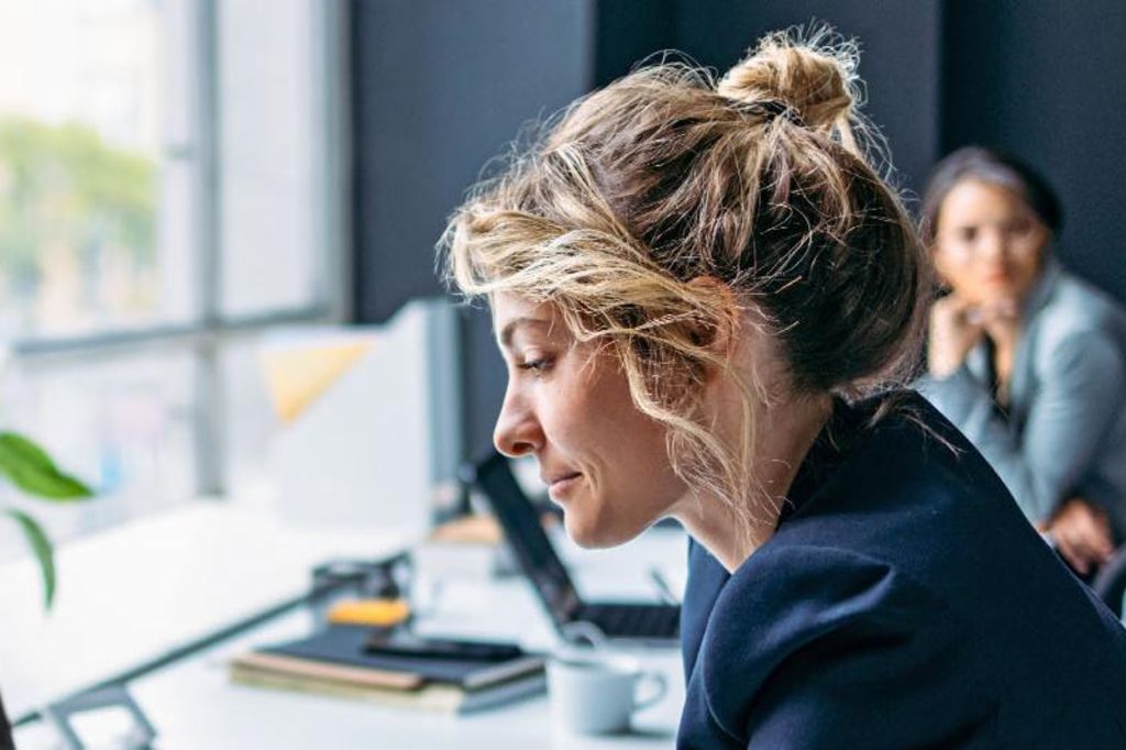 Billedet viser en kvinde, der sidder ved et skrivebord i et moderne kontor, iført en mørk blazer, mens hun fokuserer på sin bærbare computer, med en plante og nogle kontorartikler synlige på bordet, og en anden person sidder i baggrunden.