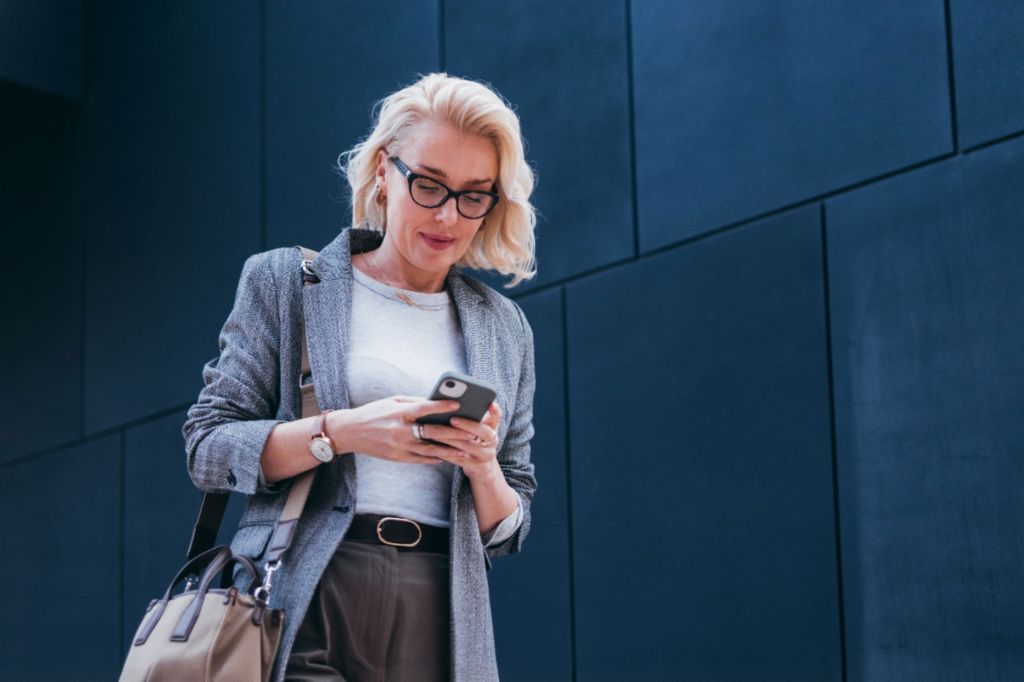 En blond forretningskvinde i en grå blazer står foran en moderne bygning og smiler, mens hun kigger på sin smartphone.