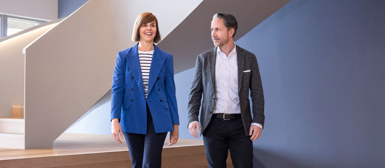 CEO'en i en grå blazer og CFO'en for EOS Gruppen i en blå blazer går gennem et moderne kontor. Rummet har store vinduer, en buet væg og stilfulde hængende lamper.