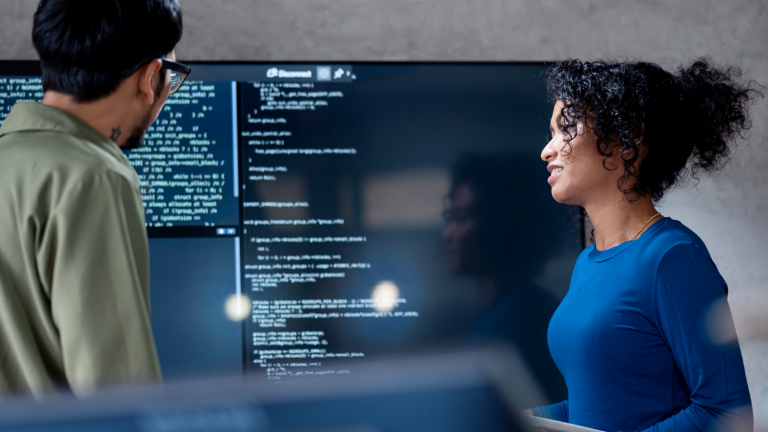 AI i inkasso: I dette billede analyserer to professionelle samarbejdende programkode til automatiserede processer på en stor skærm. Scenen illustrerer den praktiske anvendelse af Kunstig Intelligens (AI) og Robotisk Procesautomatisering (RPA) i gældshåndtering – et eksempel på digital transformation i fordringsadministration.