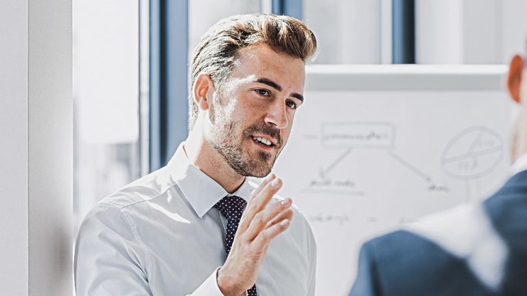 Man ser en mand i forretningstøj forklare noget med et venligt og engageret udtryk. Foran ham står en anden person, som tydeligt lytter til ham. I baggrunden ses en whiteboardtavle med en skitse.