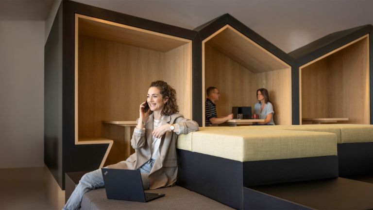 En smilende person sidder i en moderne kontorlounge, taler i telefon og arbejder på en bærbar computer. I baggrunden arbejder to andre personer.