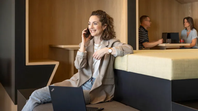 En smilende person sidder i en moderne kontorlounge, taler i telefon og arbejder på en bærbar computer. I baggrunden arbejder to andre personer.