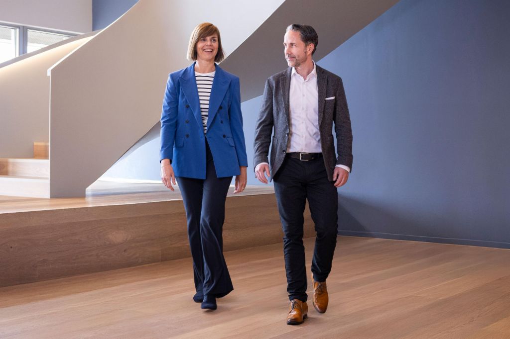 CEO'en i en grå blazer og CFO'en for EOS Gruppen i en blå blazer går gennem et moderne kontor. Rummet har store vinduer, en buet væg og stilfulde hængende lamper.