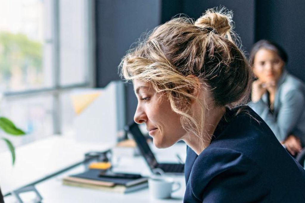 Billedet viser en kvinde, der sidder ved et skrivebord i et moderne kontor, iført en mørk blazer, mens hun fokuserer på sin bærbare computer, med en plante og nogle kontorartikler synlige på bordet, og en anden person sidder i baggrunden.