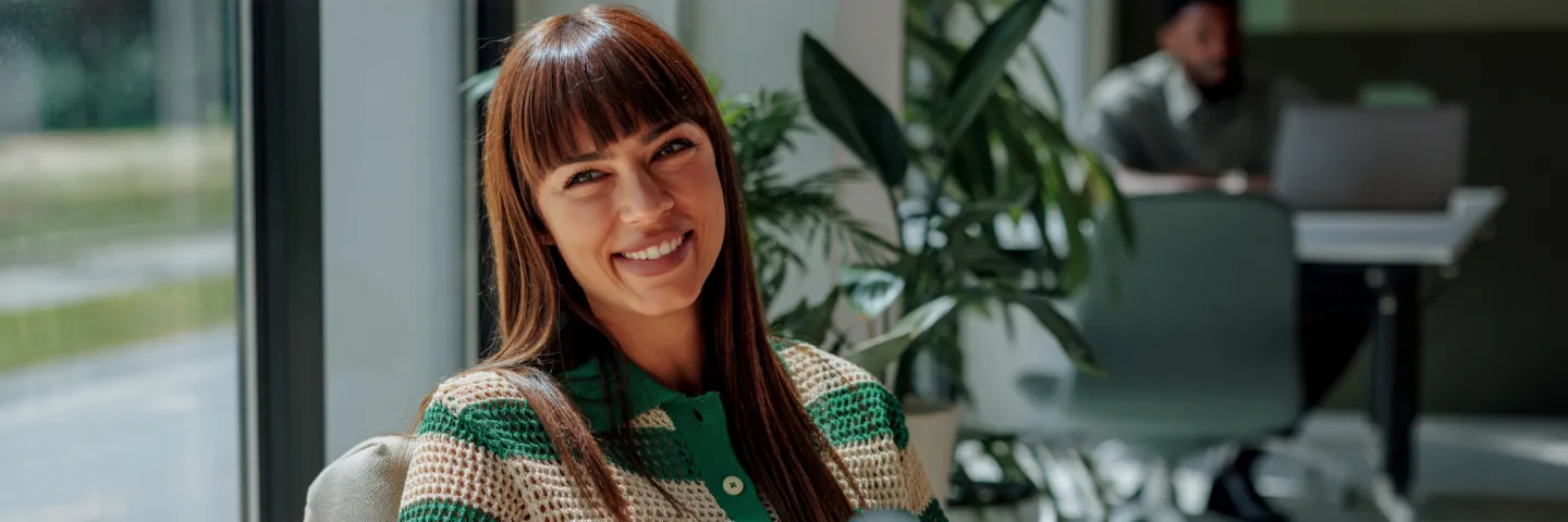 En smilende kvinde med mørkt hår sidder komfortabelt i et lyst, moderne kontor og ser varmt ind i kameraet. I baggrunden ses andre personer og store vinduer.