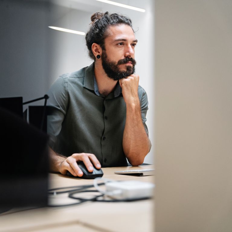 En skægget mand med langt hår bundet i en knold sidder ved en computer, fuldt opslugt af sit arbejde.