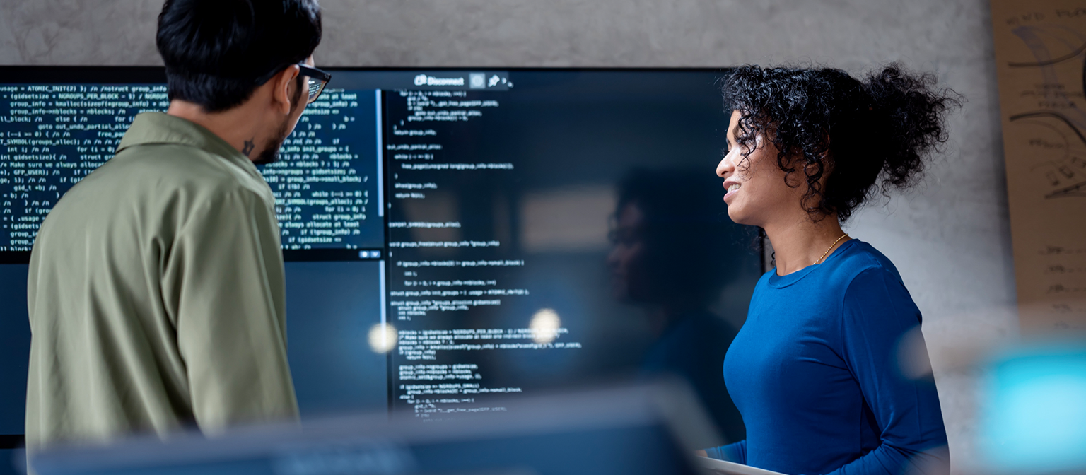 AI i inkasso: I dette billede analyserer to professionelle samarbejdende programkode til automatiserede processer på en stor skærm. Scenen illustrerer den praktiske anvendelse af Kunstig Intelligens (AI) og Robotisk Procesautomatisering (RPA) i gældshåndtering – et eksempel på digital transformation i fordringsadministration.