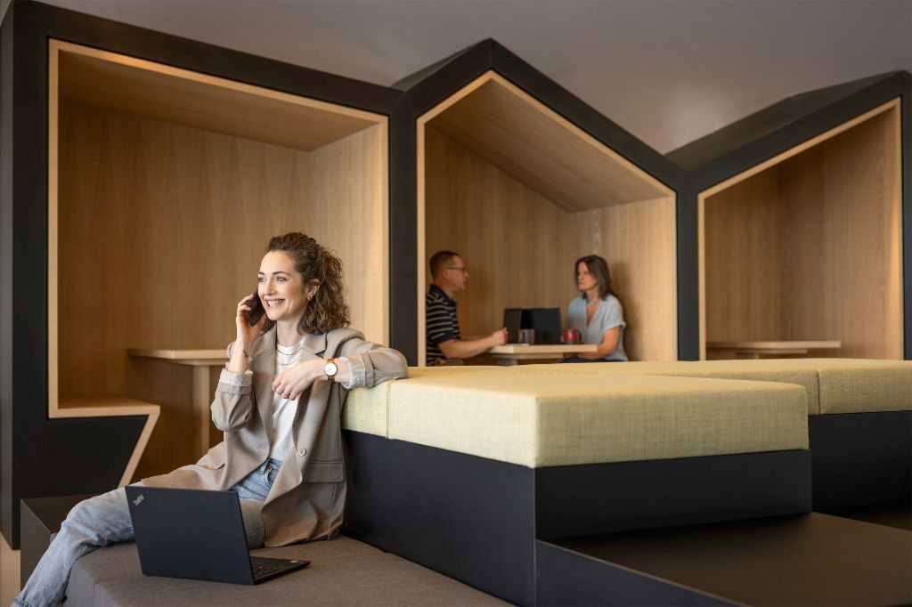 En smilende person sidder i en moderne kontorlounge, taler i telefon og arbejder på en bærbar computer. I baggrunden arbejder to andre personer.